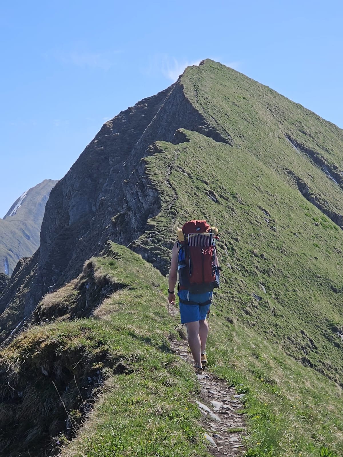 Wanderin mit grossem Rucksack auf dem schmalen grasigen Grat, beidseitig fallen die Hänge steil ab